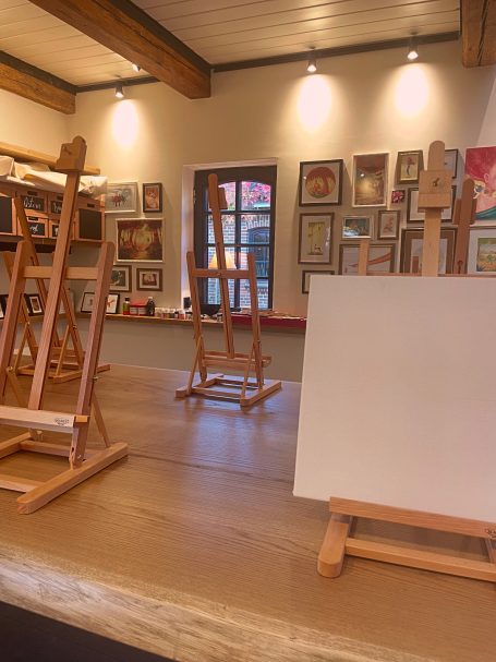 Ein Raum mit Staffeleien und einer leeren Leinwand, umgeben von bunten Kunstwerken.